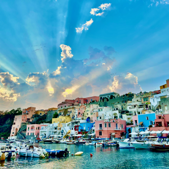 Colorful hillside houses and boats at a harbor under a dramatic sky with sunrays.