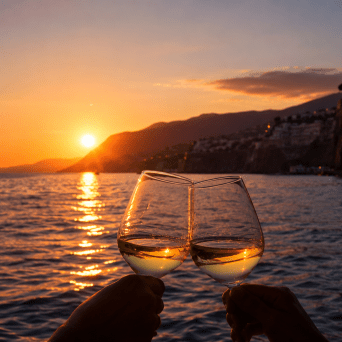 Two wine glasses clinking at sunset by the sea with mountains in the background.