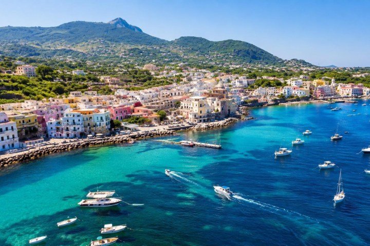 Coastal town with colorful buildings, boats in blue waters, and hills in the background.