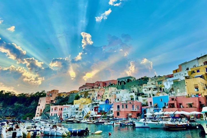Colorful seaside houses with boats in harbor under a vibrant sky with sunrays breaking through clouds.