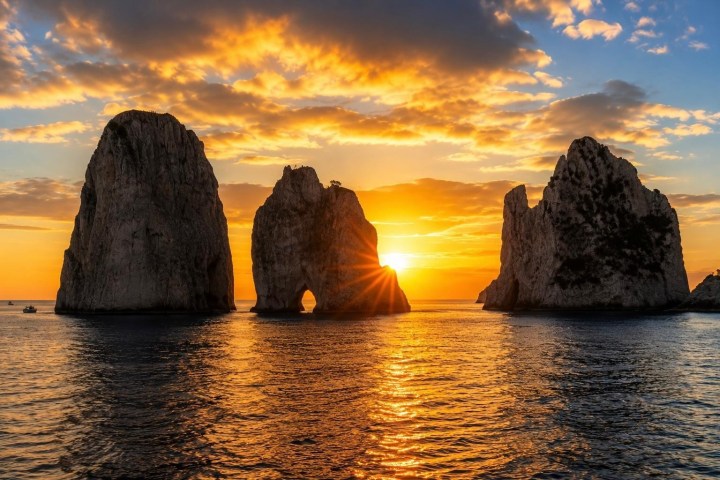 Sunset over ocean with three large rock formations and golden clouds.