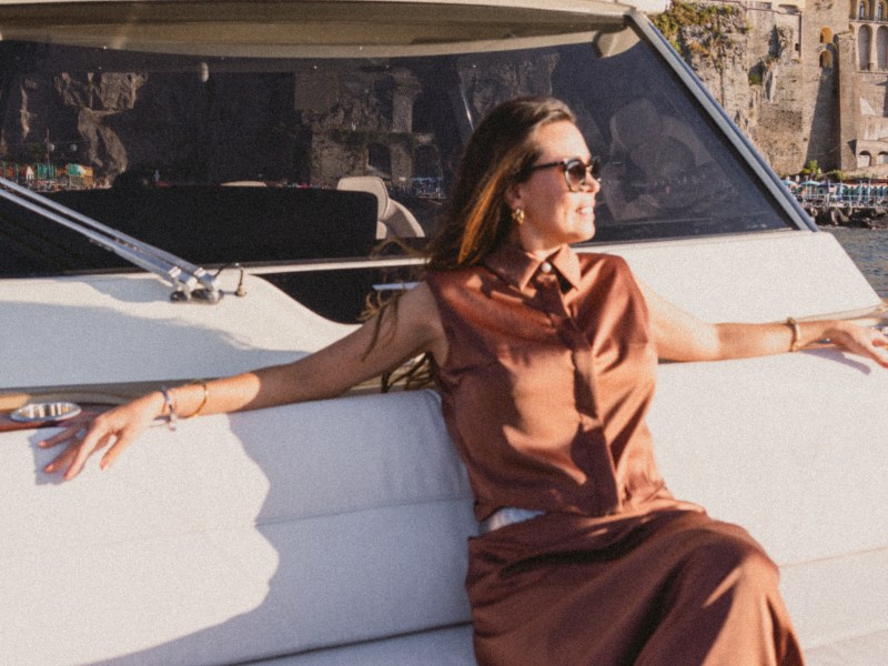 Woman in sunglasses relaxing on a boat with coastal buildings in the background.